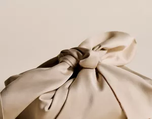 Nahaufnahme der Hand einer Person, die einen Knoten an einem beigen Furoshiki-Tuch bindet, vor einem beigen Hintergrund